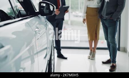 Wenig Aufnahme von wohlhabenden Menschen, jungen Paaren, die im Händlerbetrieb ein neues Auto kaufen und mit einem hilfsbereiten Manager im Anzug sprechen. Glänzendes Luxusauto ist im Vordergrund, großes Fenster im Hintergrund. Stockfoto
