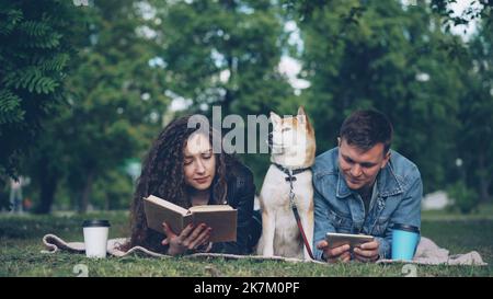 Glückliches junges Paar ruht im Park, Mann benutzt Smartphone, während Frau Buch liest, ihr Hund sitzt zwischen ihnen auf einer Decke. Die Menschen reden und lächeln. Stockfoto