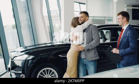 Der junge bärtige Mann kauft ein Auto für seine attraktive Frau, sie küsst und umarmt ihn. Stockfoto