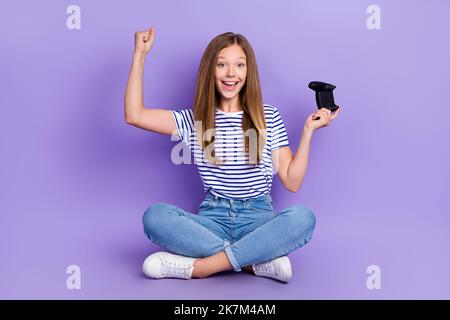 In voller Länge Foto von ekstatischen Mädchen tragen gestreifte T-Shirt sitzen auf dem Boden Schrei ja halten Joystick gewinnen Spiel isoliert auf lila Farbe Hintergrund Stockfoto