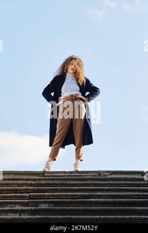 Seriöse Frau in blauem Wolltrenchmantel steht an sonnigen Herbsttag auf Treppen Stockfoto