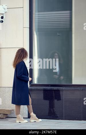 Nicht erkennbare Frau im Mantel geht den Bürgersteig entlang und schaut auf ihre Spiegelung in den Fenstern Stockfoto