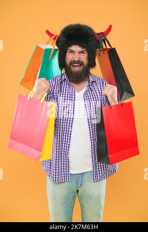 Tolle Anschaffung. Bärtiger Mann, der mit einem persönlichen Kauf lächelte. Glücklicher Hipster in Stierhörnern Hut hält seinen Kauf in Paperbags. Mode kaufen Stockfoto
