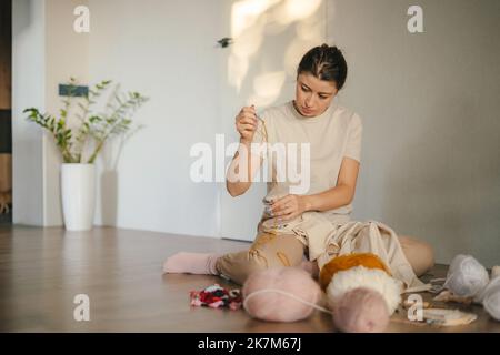 Porträt einer konzentrierten Frau, die von Hand im Kreis ein Muster auf hellem Material stickt und im Raum auf dem Boden sitzt. Das Konzept Stockfoto