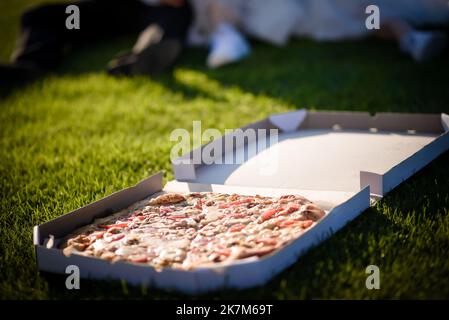 Ein fröhliches Paar junger Leute, die Spaß haben, auf dem Boden zu sitzen, Wein zu trinken und Pizza zu beißen - ein Lifestyle-Konzept für romantische Leute Stockfoto