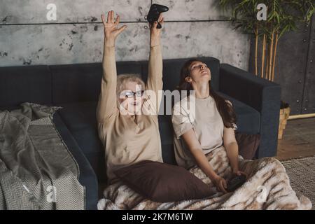 Kaukasische ältere Frau feiert, dass sie die Hände hochhält, während sie mit ihrer Tochter zu Hause Videospiele spielt. Spiel- und Unterhaltungskonzept. Glücklich Stockfoto