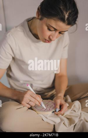 Schöne kaukasische Frau, die das Schema auf die Leinwand zeichnet, die sie besticken wird. Textilgewebe. Stickerei in Frauenhänden. Stockfoto