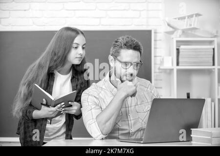 Aufmerksames Kind und Vater sitzen im Klassenzimmer mit Copybook und Computer an der Tafel, Schularbeiten Stockfoto