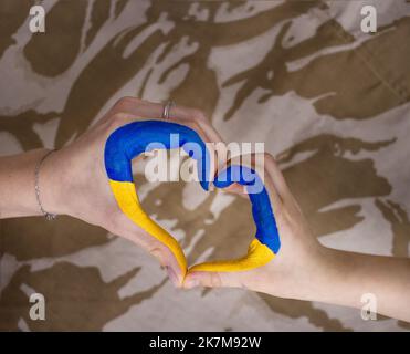 Die Hand von Frau und Kind wurde in Form eines Herzens auf einem Camouflage-Hintergrund gefaltet. Familie, Patriotismus, Einheit, Unterstützung. Russlands Invasion der Ukraine, c Stockfoto