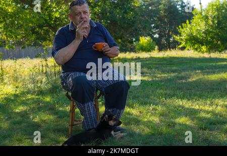 Außenportrait des ukrainischen Senioren, der im Sommergarten mit einer Teekappe sitzt, während sein kleiner Welpe in der Nähe der Füße des Meisters sitzt Stockfoto