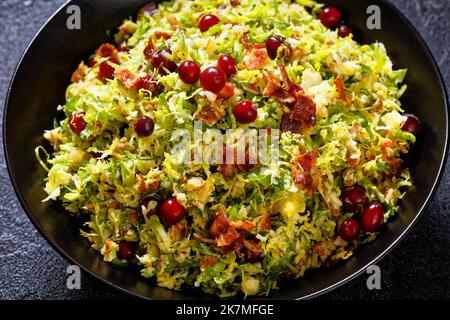 Nahaufnahme von rosenkohl mit Mandeln, knusprigem gebratenem Speck und Preiselbeeren in einer schwarzen Schüssel auf einem Betontisch Stockfoto