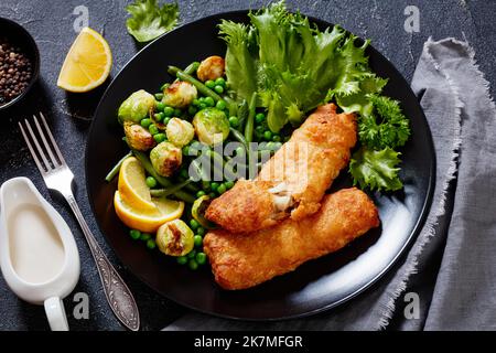 Knuspriger Bierteig Kabeljau Fischfilet mit gebratenen rosenkohl, grünen Bohnen, grünen Erbsen und frischem Salat auf schwarzem Teller auf Betontisch Stockfoto