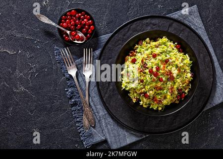 rosenkohl mit Mandeln, knusprig gebratenem Speck, geriebenem Parmesan und Preiselbeeren in schwarzer Schale auf Betontisch, horizontale Ansicht von Stockfoto