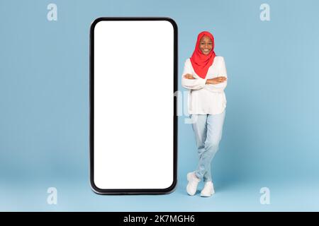 Fröhliche schwarze Frau in Hijab posiert in der Nähe von riesigen Handy mit leerem Bildschirm, bietet neueste mobile App oder coole Website Stockfoto