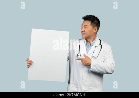 Lächelnder koreanischer Mann mittleren Alters in weißem Mantel mit Stethoskop zeigt den Finger auf ein Banner mit Kopierraum Stockfoto