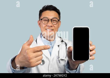 Frohes chinesisches Mann im mittleren Alter Therapeut in weißem Mantel und Brille zeigt auf Smartphone mit leerem Bildschirm Stockfoto