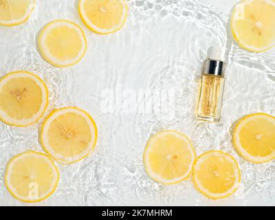 Luxuriöses kosmetisches Peeling oder Serum in Glasflasche mit Pipette in sauberem, transparentem Wasser mit Zitronenscheiben auf klarem Wasserhintergrund. Wasser spritzt im Sonnenlicht auf die Wasseroberfläche. Flatlay-Kopierbereich Stockfoto