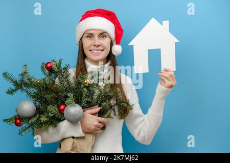 Schöne junge kaukasische Frau in Weihnachten roten Hut mit Strauß von Fichtenzweigen und Papierhaus Modell, isoliert auf blauem Hintergrund mit Kopie s Stockfoto