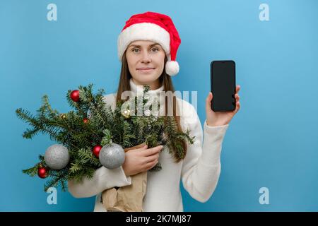 Portrait der jungen Brünette Frau trägt weißen Pullover und Weihnachtshut halten Bouquet von Fichtenzweigen verwenden Handy mit leerem Bildschirm workspa Stockfoto