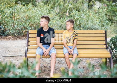 Die Brüder sitzen müde vom langen Spaziergang im Park auf der Bank. Die blonden Jungen entspannen sich auf einer Holzbank in der Nähe von Bäumen und schauen zur Seite Stockfoto