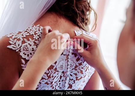 Brautmorgen, Braut trägt Kleid. Der Bräutigam hilft dem Verlobten beim Anziehen und passt die Knöpfe am Hochzeitskleid an, Rückansicht. Nahaufnahme, Ernte Stockfoto