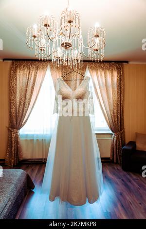 Das perfekte Hochzeitskleid mit einem vollen Rock auf einem Kleiderbügel in das Zimmer der Braut mit blauen Vorhängen Stockfoto