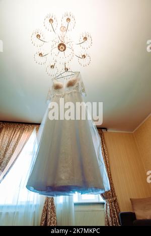 Das perfekte Hochzeitskleid mit einem vollen Rock auf einem Kleiderbügel in das Zimmer der Braut mit blauen Vorhängen Stockfoto