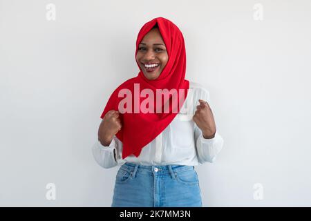 Fröhliche afroamerikanische Frau in Hijab gestikuliert ja, feiert Erfolg über weißen Studio Wand Hintergrund Stockfoto