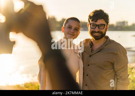 Vintage-Kamera. Schwarze männliche Fotografen POV. Verschwommener Vordergrund. Kaukasisches Paar - kahle Frau und bärtiger Mann - in lässiger Kleidung lächelnd und posiert während des Fotoshootings. Goldene Stunde. Fluss im Hintergrund. Hochwertige Fotos Stockfoto