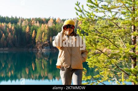 Junge Frau in gelbem Hut schaut durch ein Fernglas auf Vögel am See gegen den Herbstwald Vogelbeobachtung, Zoologie, Ökologie. Forschung in der Natur, Beobachtung Stockfoto