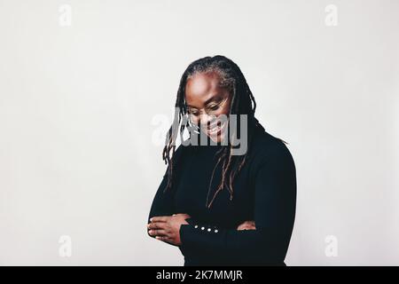 Glückliche Frau mit Dreadlocks lächelt fröhlich, während sie eine Brille trägt. Professionelle reife Frau steht vor einem grauen Hintergrund mit ihren Armen cr Stockfoto