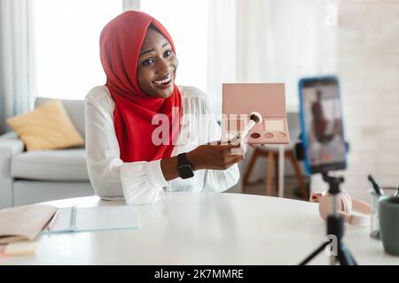 Glückliche schwarze muslimische Bloggerin zeigt Make-up-Produkt auf Handy-Kamera, macht Video mit Kosmetik zu Hause Stockfoto