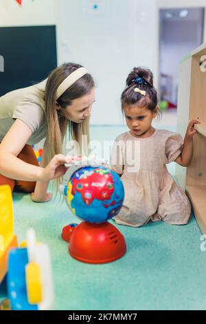 Blonde Lehrerin berührt die kleine Plastikkugel und spricht mit einem kleinen Mädchen im Kindergarten, vertikale Aufnahme. Hochwertige Fotos Stockfoto