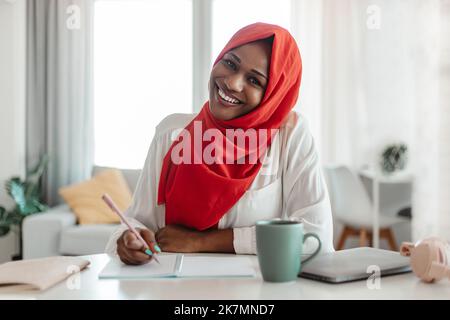 Notieren Sie sich das Konzept. Glückliche schwarze muslimische Frau, die mit einem Laptop am Schreibtisch sitzt und einen Brief in einem Papierbuch schreibt Stockfoto