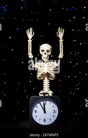 Ein menschliches Skelett winkt mit der Hand und begrüßt einen schwarzen Wecker auf einem schwarzen Hintergrund, vertikalem Rahmen, Bokeh. „Day of the Dead“-Konzept Stockfoto