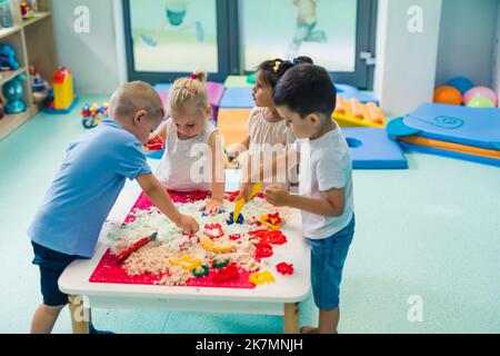 Vier Kinder, die an dem kleinen Tisch stehen und Figuren in den kinetischen Sand legen, entwickeln kreative Spiele von Vorschulkindern im Hintergrund Indoor-Spielzeuge. Hochwertige Fotos Stockfoto