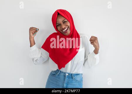 Sieg und Glück. Überglücklich schwarze muslimische Frau gestikulierte ja, schüttelte die Fäuste und feierte den Erfolg über der weißen Wand Stockfoto