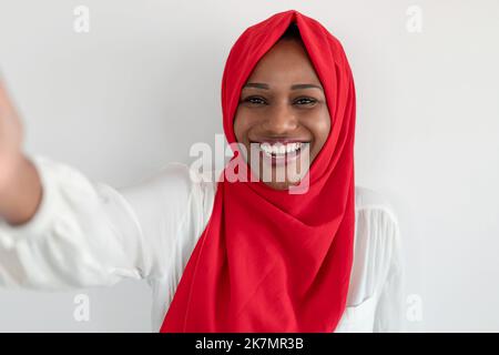 Glückliche schwarze muslimische Bloggerin macht Selfie lächelnd vor der Kamera über weißem Wandhintergrund, trägt roten Hijab Stockfoto