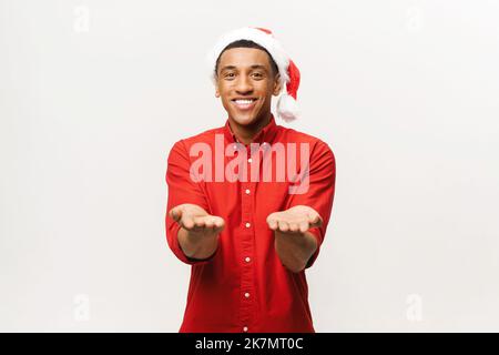 afroamerikanischer Kerl in weihnachtsmütze und rotem Hemd zieht seine Hände isoliert auf weißem Hintergrund zu dir, Kerl, der ein Geschenk gibt oder erhält, hält leeren Kopierraum über seinen Handflächen Stockfoto