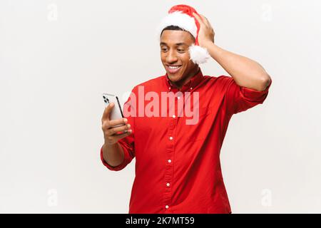 Wunderte sich glücklich jungen Mann trägt santa Hut Blick auf Smartphone und halten Kopf überrascht, isoliert auf weißem Hintergrund. Afrikanischer Mann erhielt Neujahrsgeschenk, Party-Einladung. Werbekonzept Stockfoto