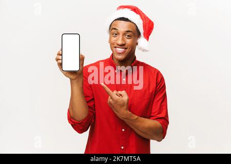 Fröhlicher afroamerikanischer junger Mann mit weihnachtsmann-Hut zeigt auf den leeren Telefonbildschirm, präsentiert Urlaubsangebote, wirbt für neue App auf dem Handy Stockfoto