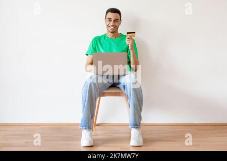 Glücklicher araber, der online per Laptop einkauft, seine Kreditkarte hält und die Kamera anlächelt, auf einem Stuhl über einer hellen Wand sitzt Stockfoto