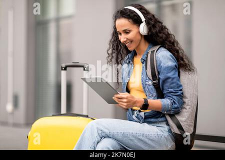 Positive junge Touristenfrau, die digitale Pads und kabellose Kopfhörer verwendet Stockfoto