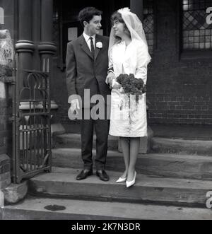 1964, historisch, ein junges, gerade verheiratetes Paar, das draußen auf den Stufen zu einer Kirche, England, Großbritannien, zusammenstand und sich gegenseitig in die Augen schaute, offensichtlich verliebt. Stockfoto