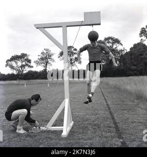 1964, historisch, Juli und draußen in einem gemähten ländlichen Feld, vor-Saison-Training für zwei Fußballer von Aylesbury vereint FC....its Steuerkurs-Training mit einem Fußball am Ende einer Schnur auf einem hohen hölzernen Apparartus, mit einem der Spieler springen, um den Ball zu führen, Buckinghamshire, England, Großbritannien. Stockfoto