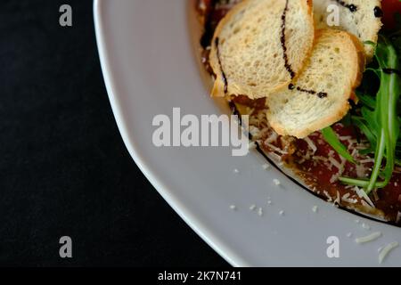 Kalte Vorspeise aus Beef-Karpacio mit Parmesan, Tomatos und Rucola auf weißem Teller mit Hardtack auf dunklem Grund. Stockfoto