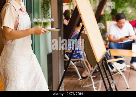 Kellnerin mit Schürze serviert dem Gast im Café einen vereisten Matcha Latte. Stockfoto