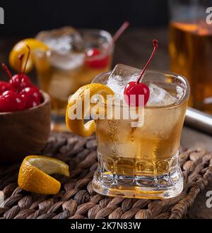 Altmodischer Cocktail mit Orangenschale und Kirsche auf einem Holztisch mit einer Schüssel mit Kirschen und einer Whiskey-Flasche Stockfoto