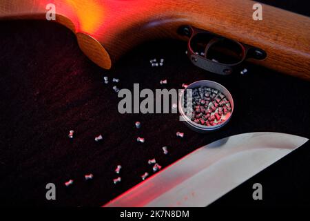 Gewehr Pistole Abzug mit Luftpellets und Messer für die Jagd auf Stoff dunklen Hintergrund mit rotem Licht. Stockfoto