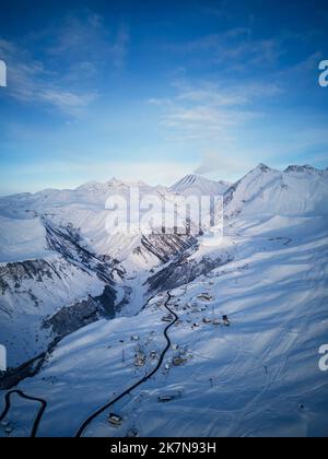 Luftaufnahme der verschneiten Bergkette bei Wintersonnenaufgang im Skigebiet. Drohne über Bergtal und Dorf mit kurviger Straße bei Sonnenuntergang. Gipfel des Kaukasus Stockfoto